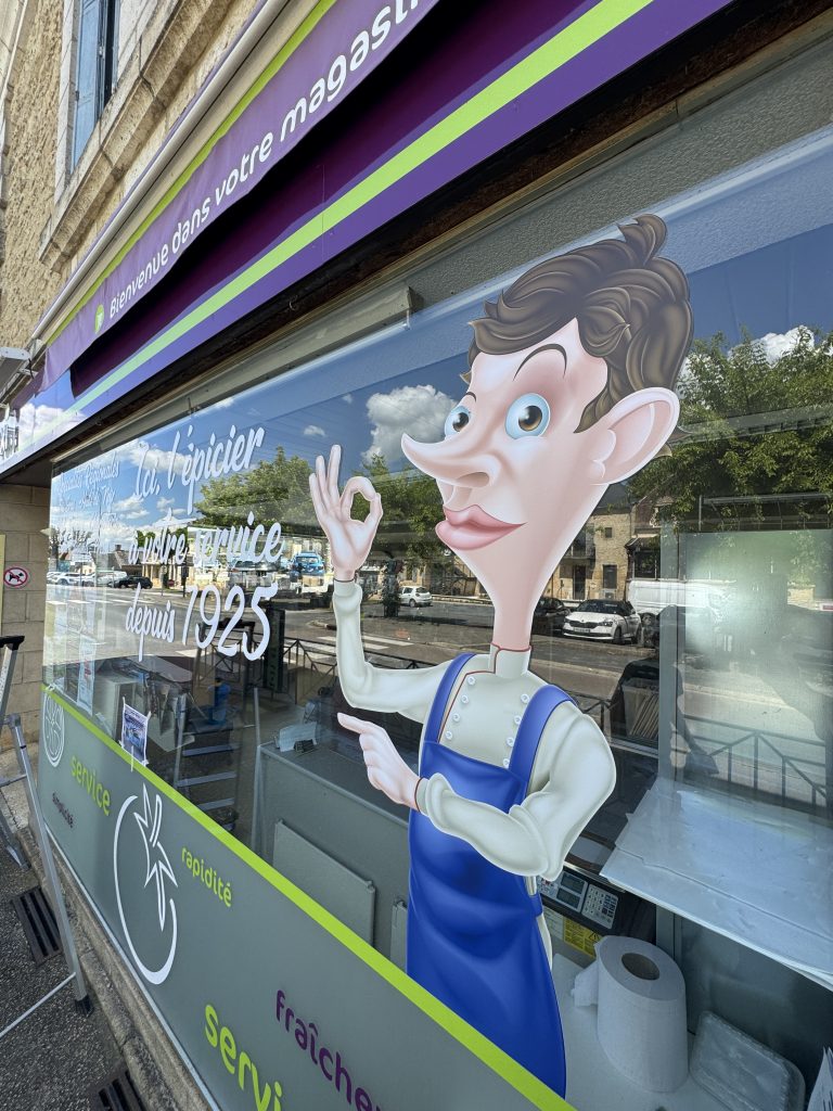 Le personnage de l’épicier découpé à sa forme et l’accroche « Ici, l’épicier à votre service depuis 1925 » sur la vitrine Proxi