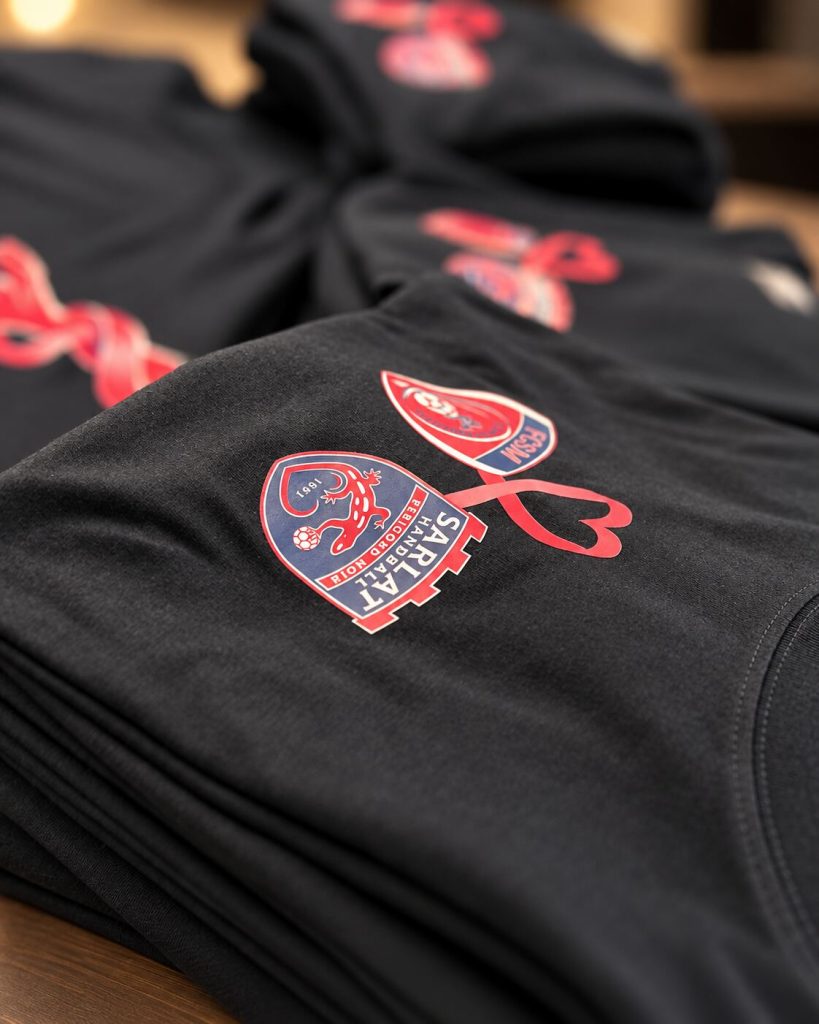 Gros plan des écussons du FCSM et de Sarlat Handball imprimés en DTF côté cœur, reliés par un ruban rose